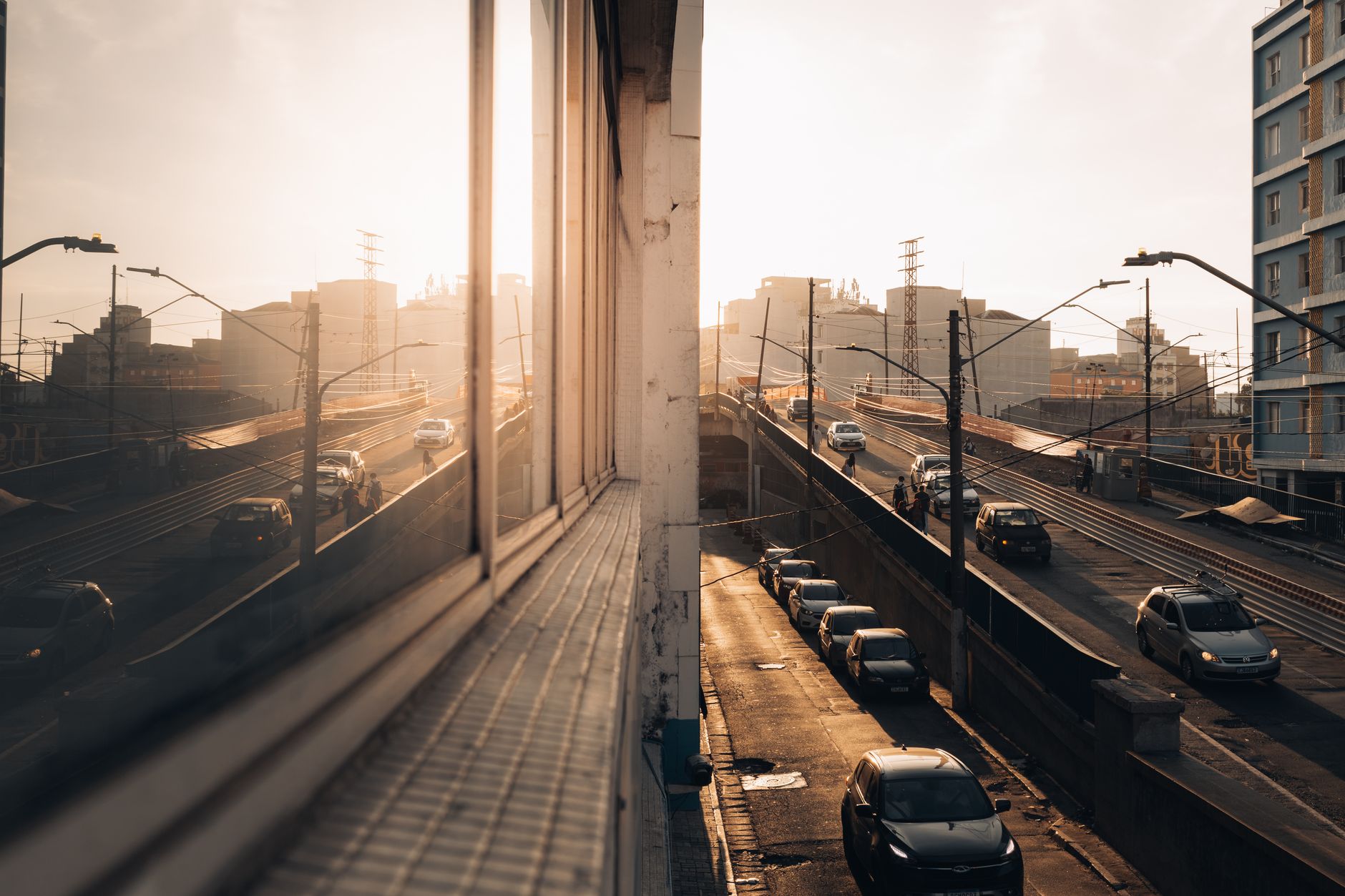 Cars drive on an urban highway during sunset, reflected in a building's windows, creating a warm atmosphere.