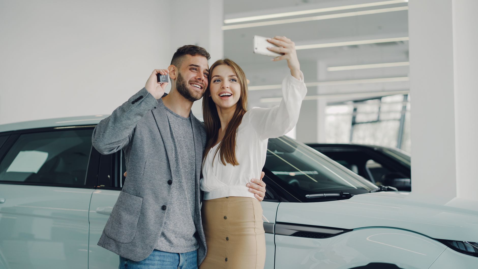 Smiling couple taking a selfie with car keys in a showroom, celebrating their new vehicle purchase.