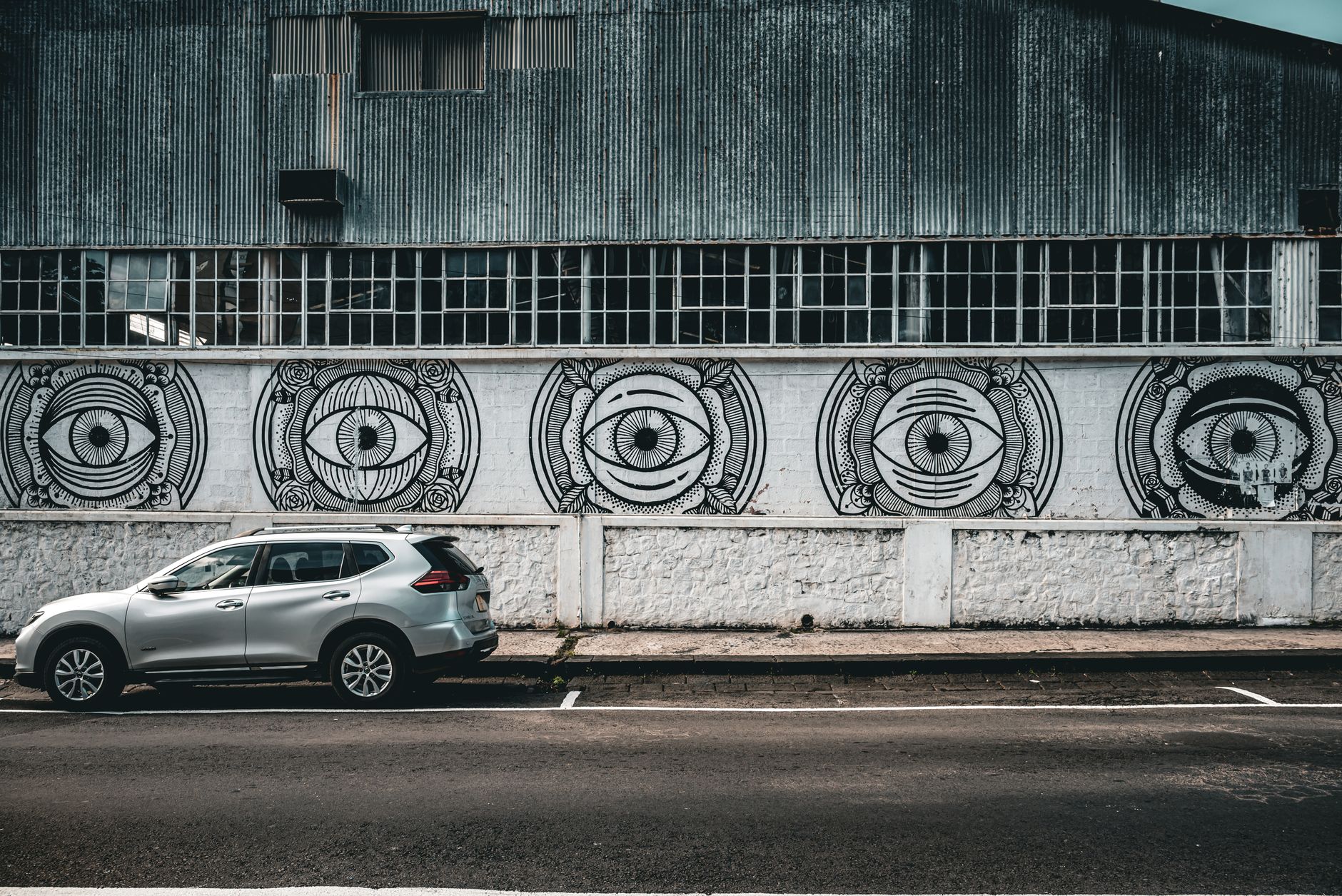 Street art mural featuring eye designs on warehouse wall with a silver SUV parked in front.