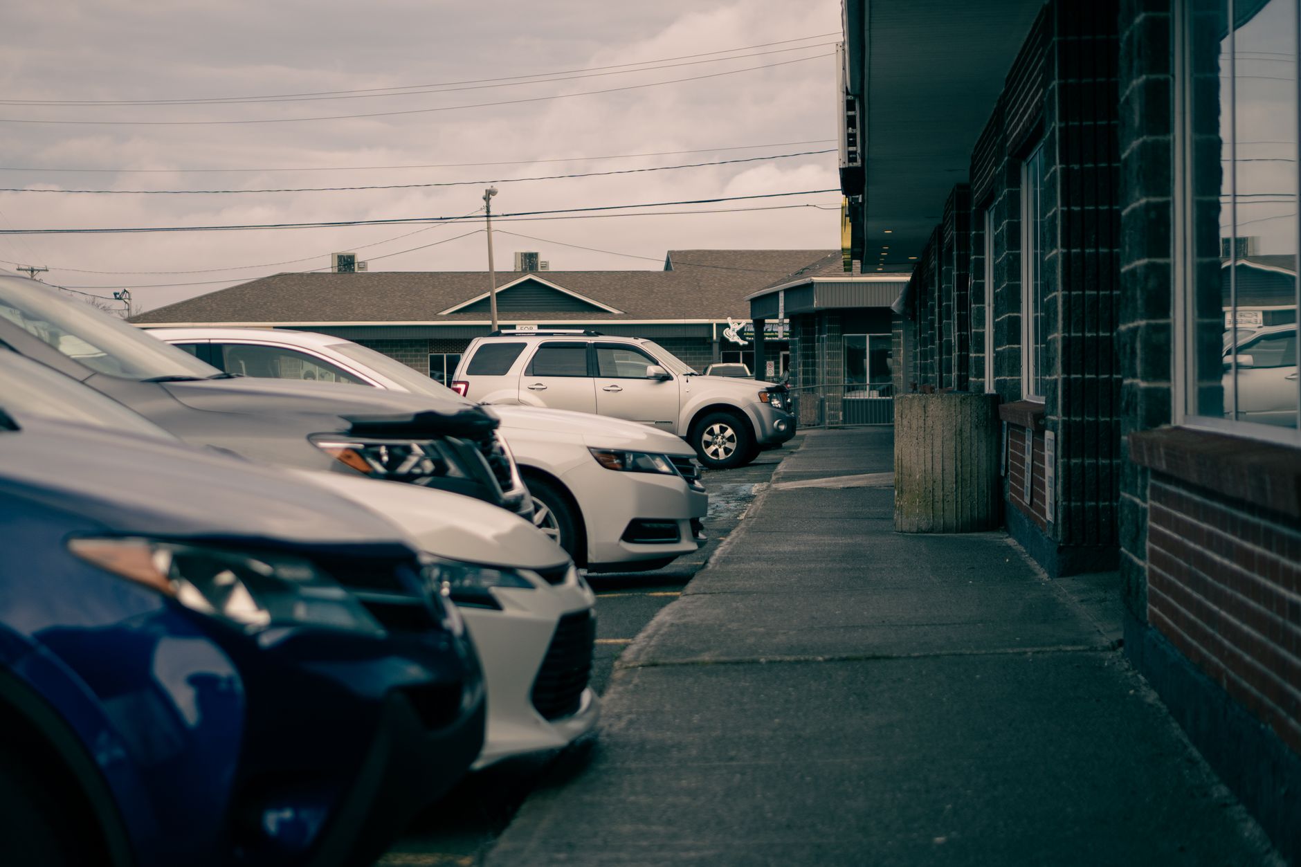 Row of contemporary automobiles parked against walkway and house facades under cloudy sky in town