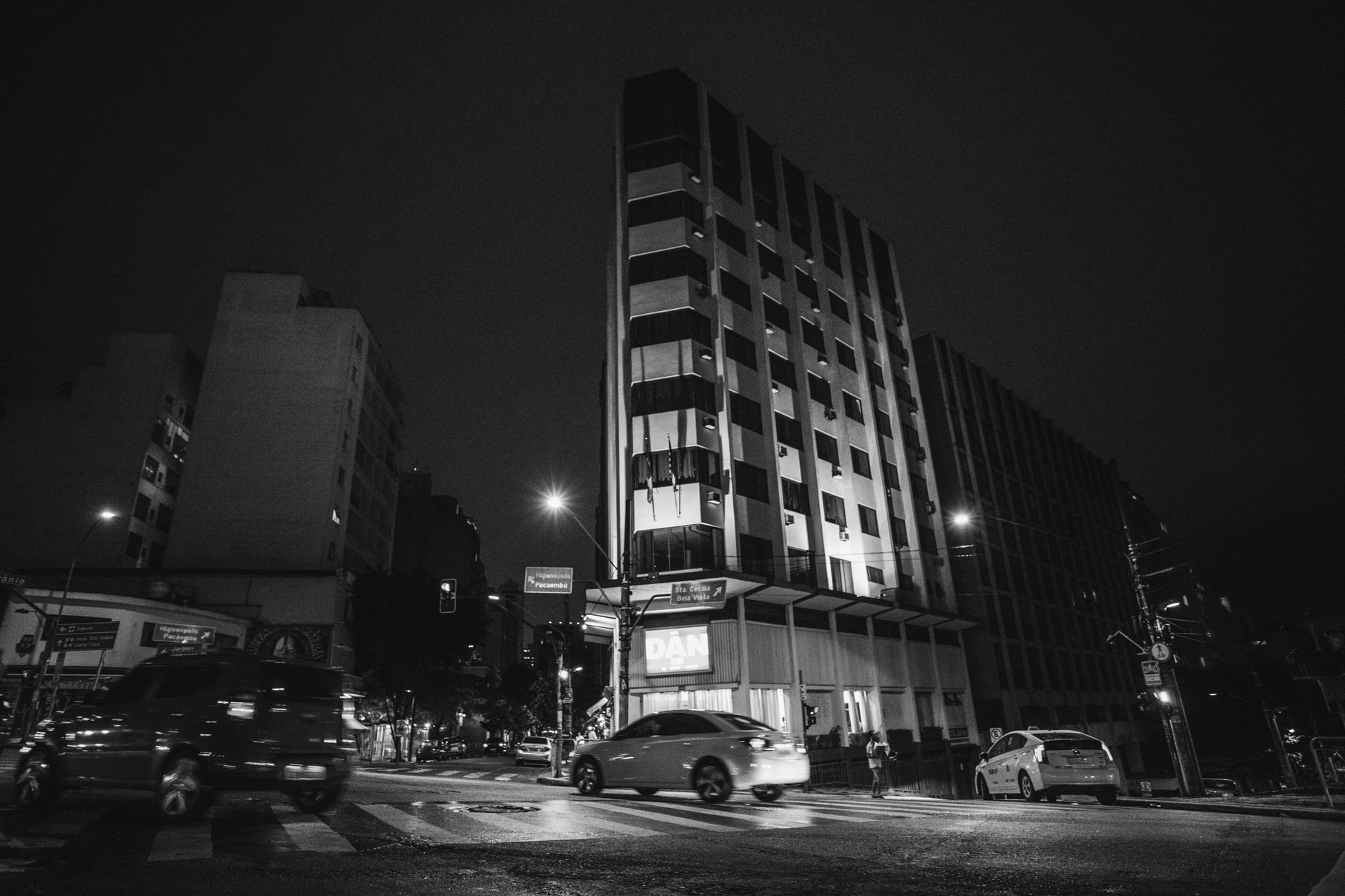 Monochrome urban scene featuring buildings and cars at a nighttime intersection.
