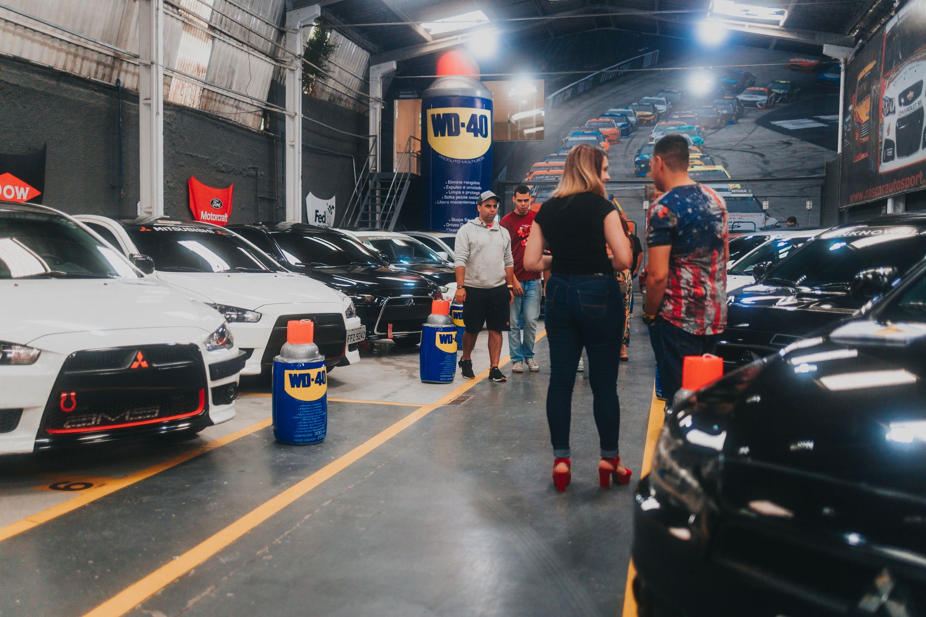 People inspecting lined-up Mitsubishi Lancer Evolutions in an indoor car show garage.