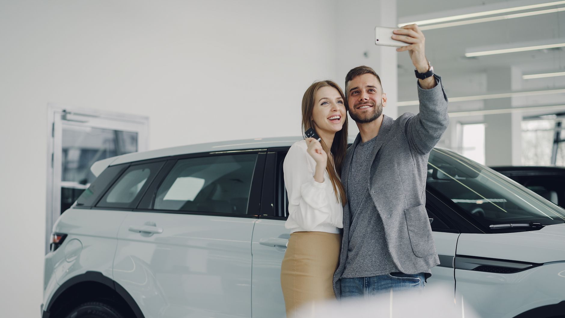 A joyful couple taking a selfie in front of their new car inside a modern showroom.