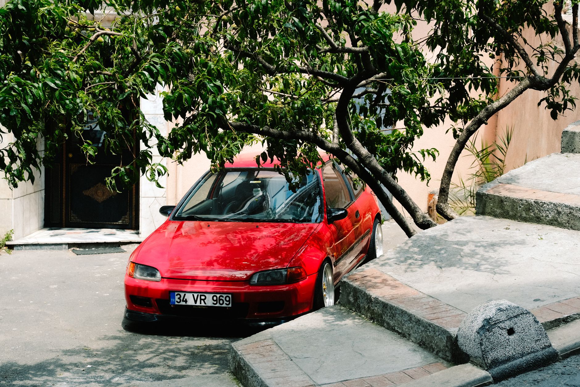A red hatchback car parked under a lush tree in an urban environment, providing contrast and shade.