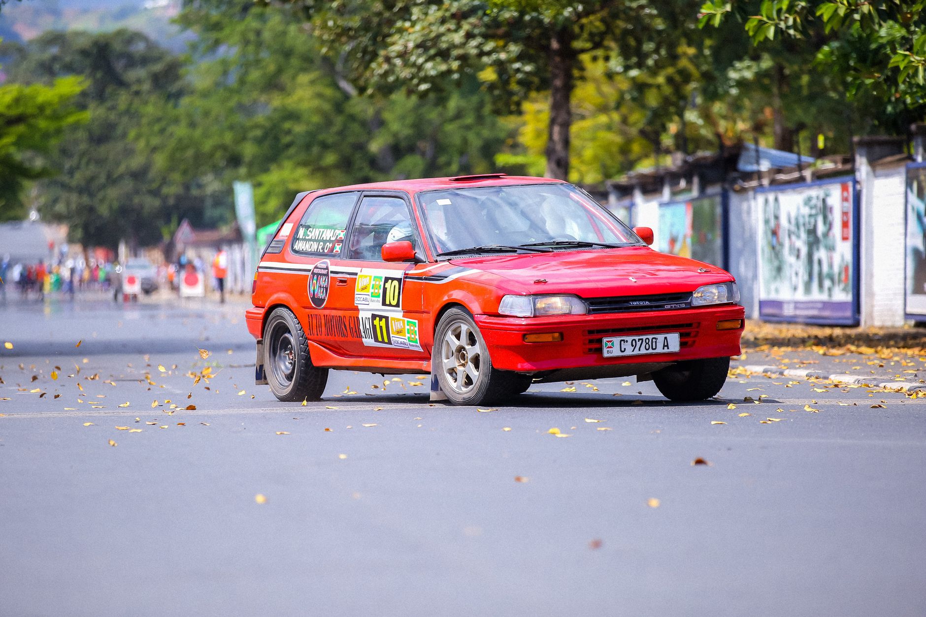 Red Toyota Corolla hatchback racing on urban streets in a classic rally event.