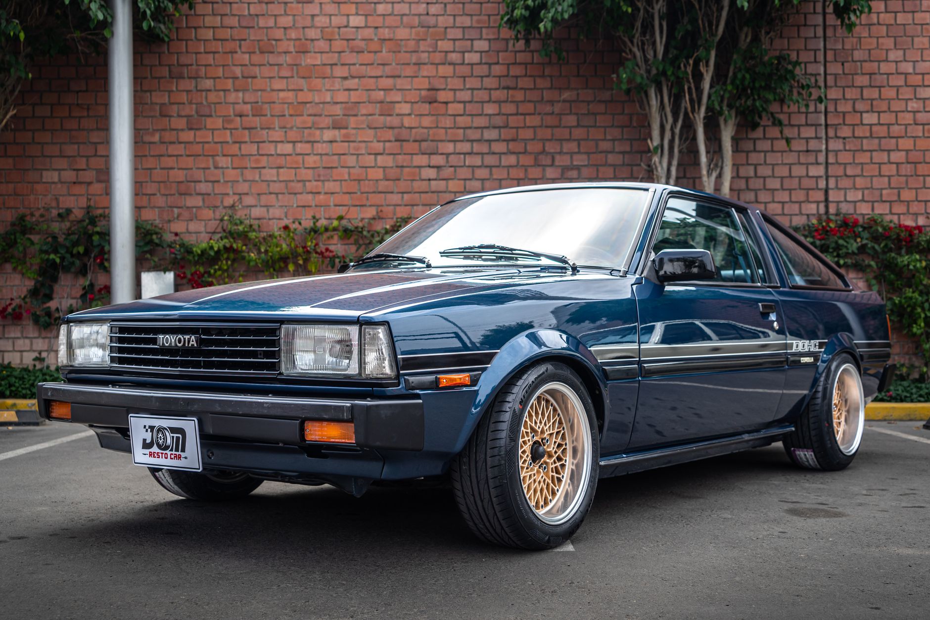 Classic Toyota AE86 Levin GT in navy blue, parked outdoors against a brick wall with greenery.