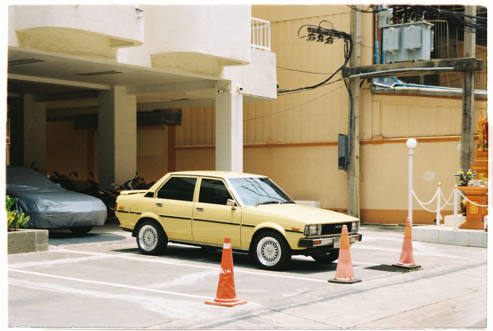 A classic yellow Toyota Corolla E70 parked in an urban setting on a sunny day, evoking a vintage feel.