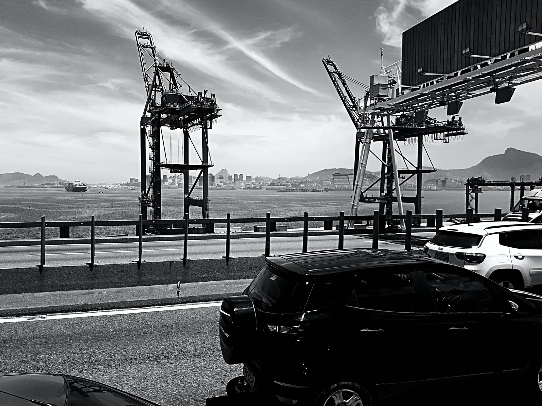 Black and white image of a bustling harbor with cranes, cars, and cityscape.