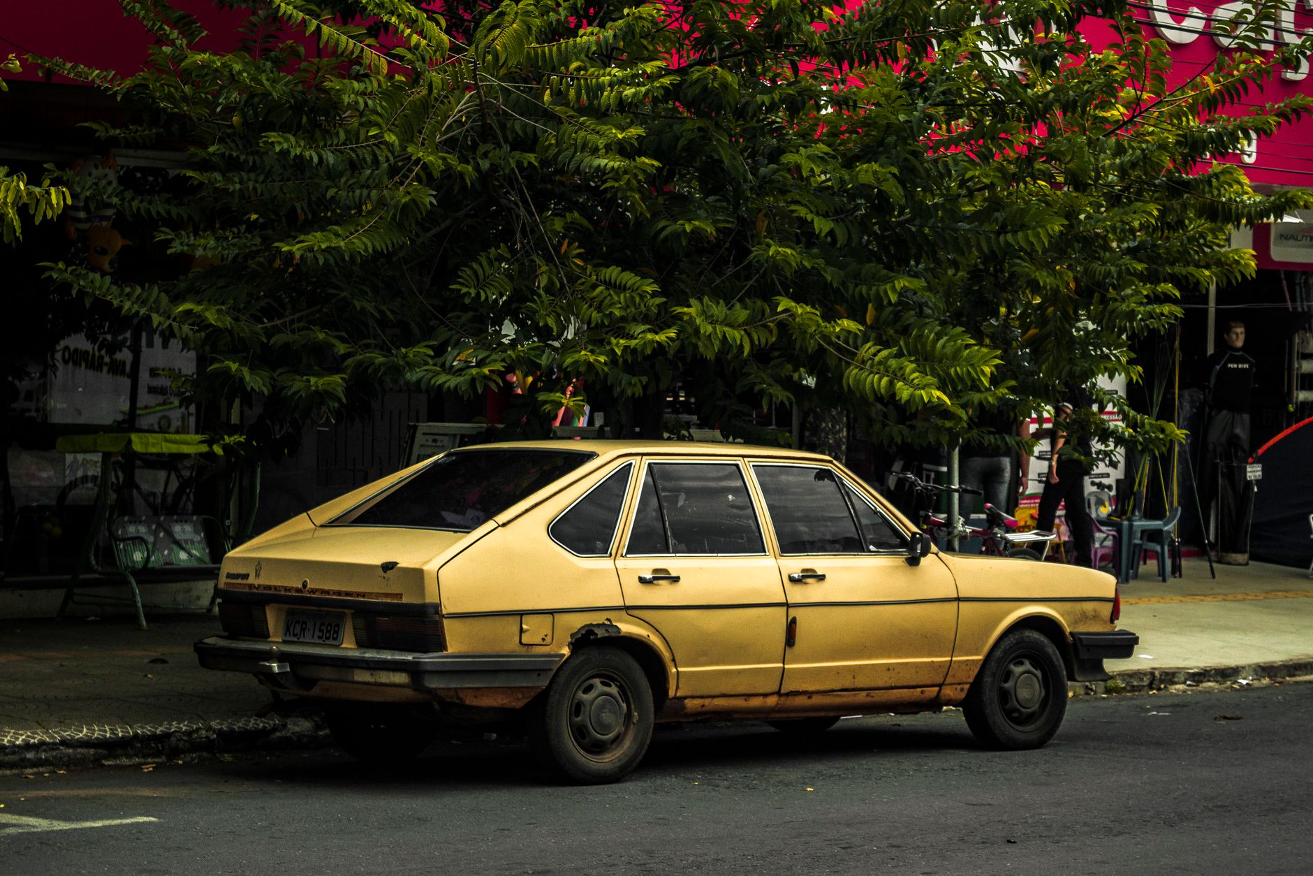 A vintage yellow car parked under a tree on a street in Goiânia, Brazil.