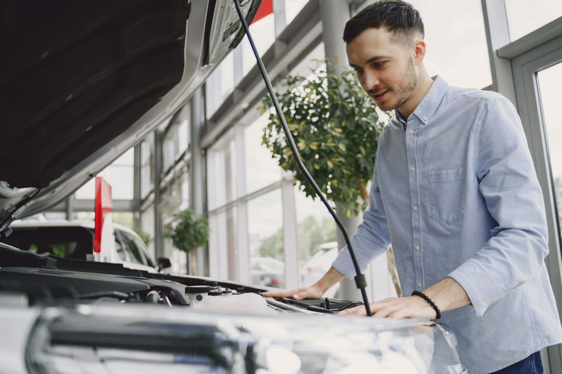 Young man examines car engine in a modern showroom, wearing a blue shirt.