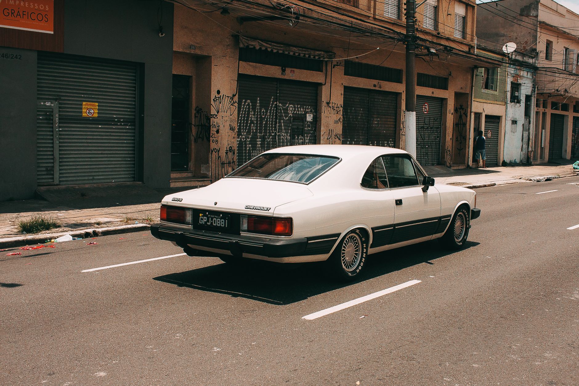 A vintage white Chevrolet Opala driving down an urban street lined with old storefronts.