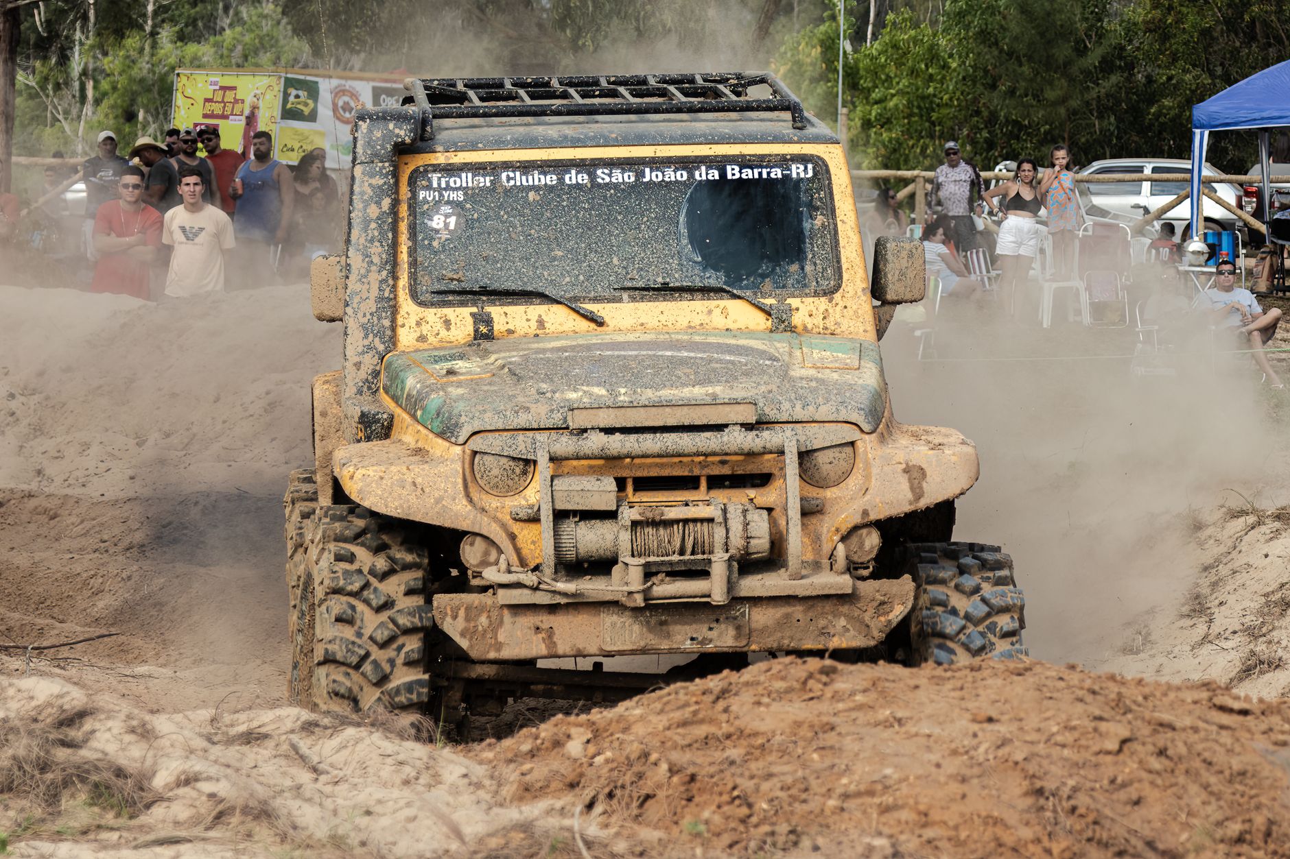 Yellow jeep in intense mud racing competition with spectators outdoors.