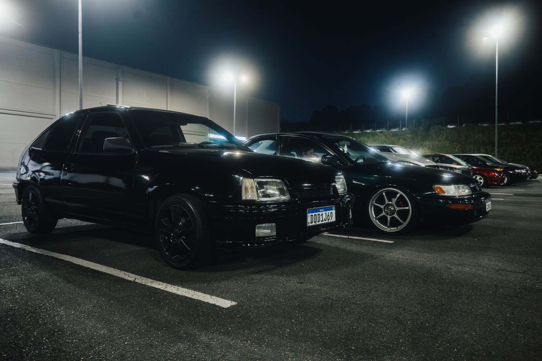Row of vintage black cars parked in a dimly lit parking lot at night under bright streetlights.