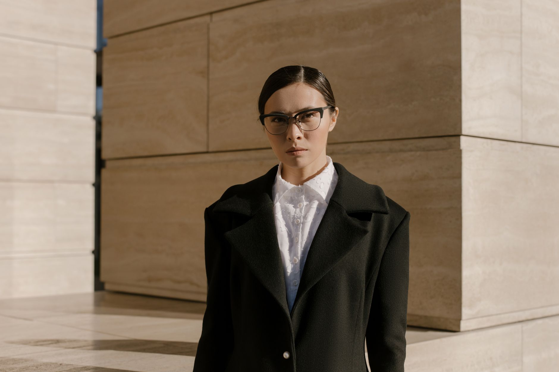 Stylish woman in a black coat and eyeglasses posed against a modern building exterior.