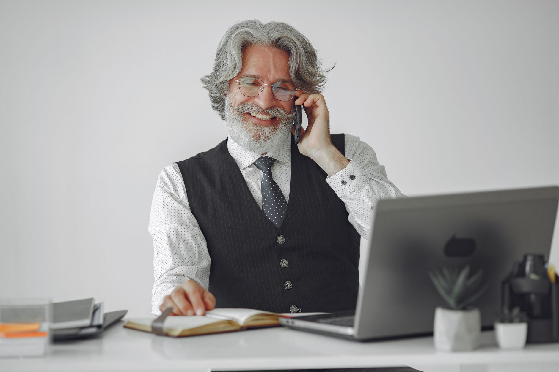 Gray-haired man in a vest multitasking with laptop and smartphone at a modern office desk.