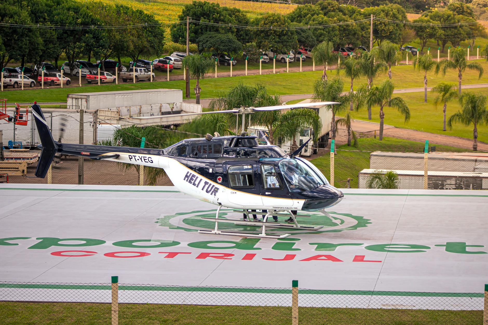 A helicopter is parked on a helipad at Expo Directa Cotrijal surrounded by palm trees and parked cars.