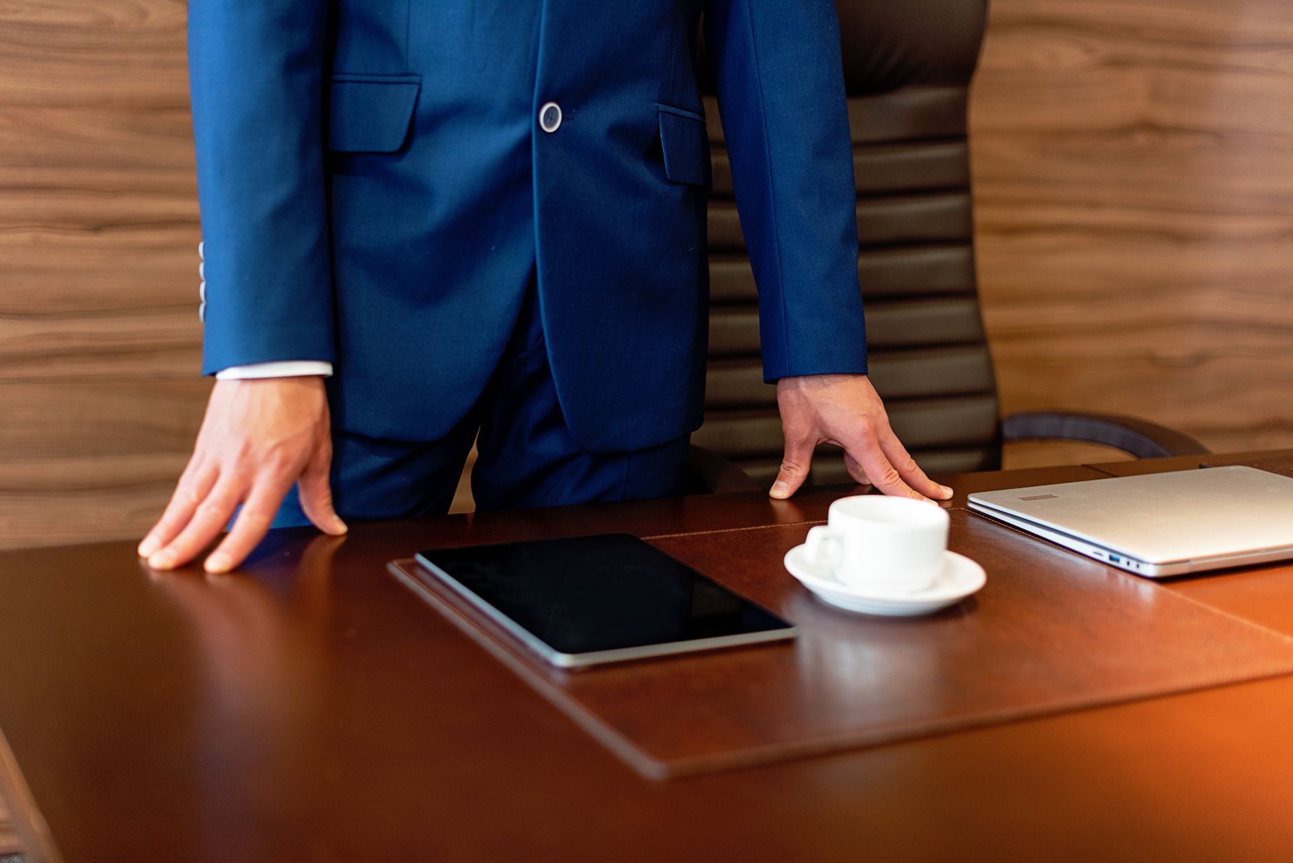 Professional businessman in blue suit standing by a desk with a tablet and coffee cup.