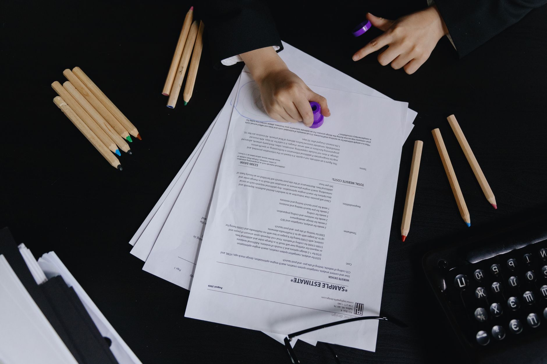 Hands working with documents and colored pencils on a black desk view.