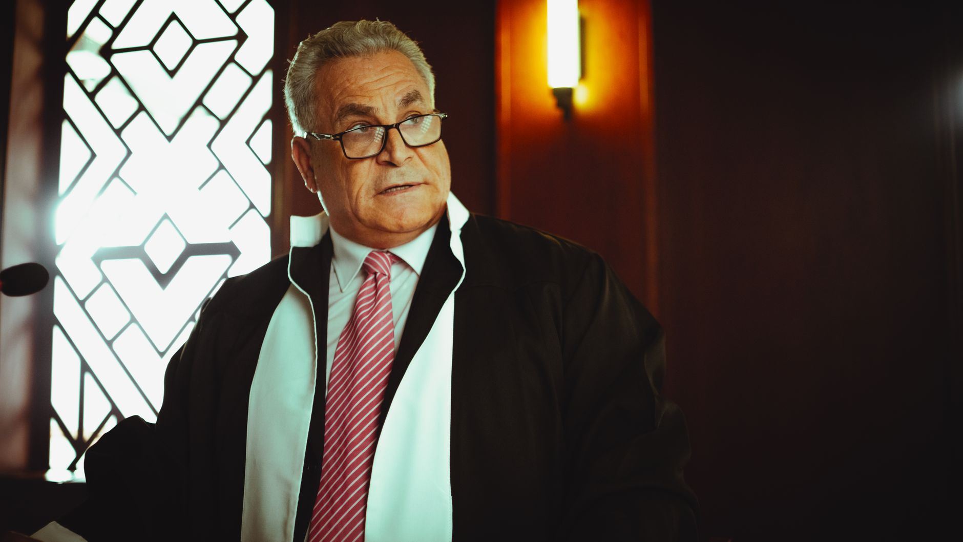 A male judge in a courtroom in Baghdad wearing a robe and tie, in a contemplative pose.