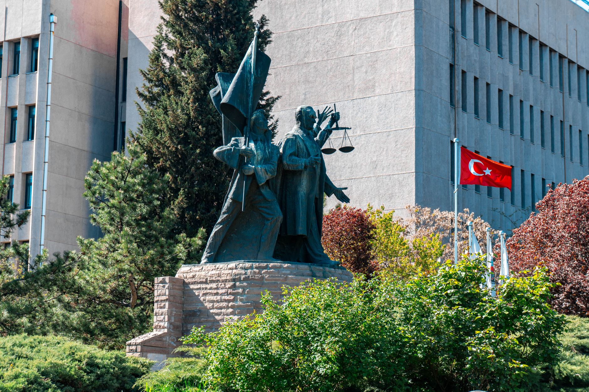 Monument before a government building in Ankara, Turkey, featuring flags and greenery.