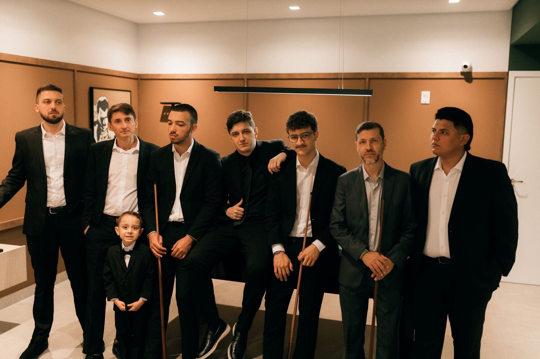 Seven men and a boy in suits gathering around a pool table indoors.