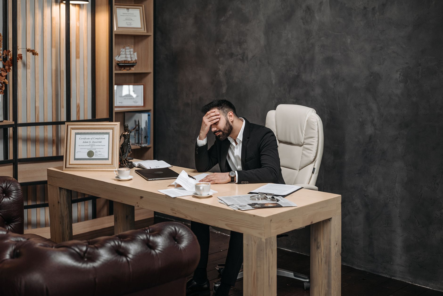 Businessman in a black suit looking stressed at an office desk, holding his head with papers scattered around.