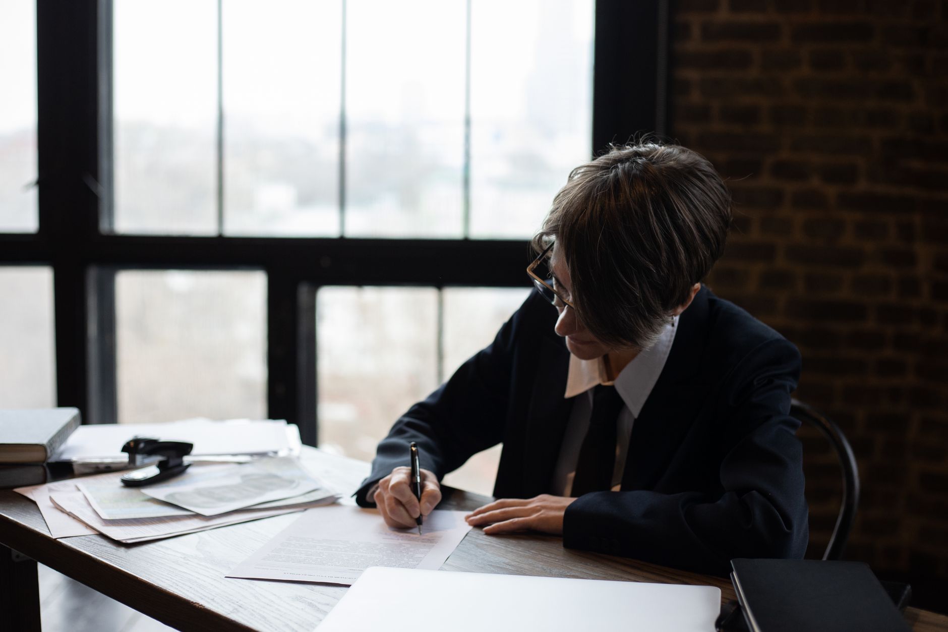 Business professional in formal attire writing at a desk in an office setting with large windows.