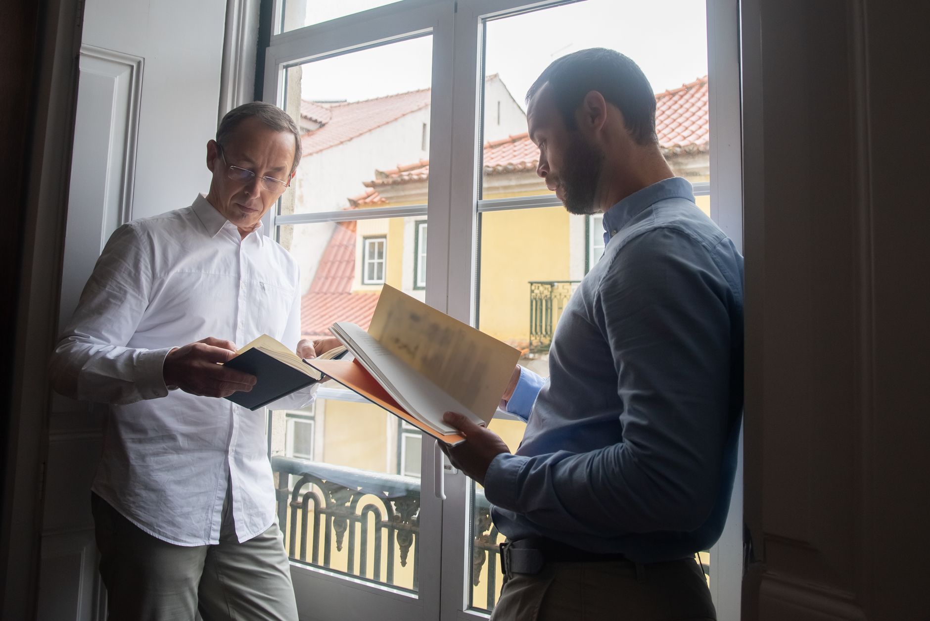 Two men standing by a window, reading and enjoying leisure time in Lisbon.