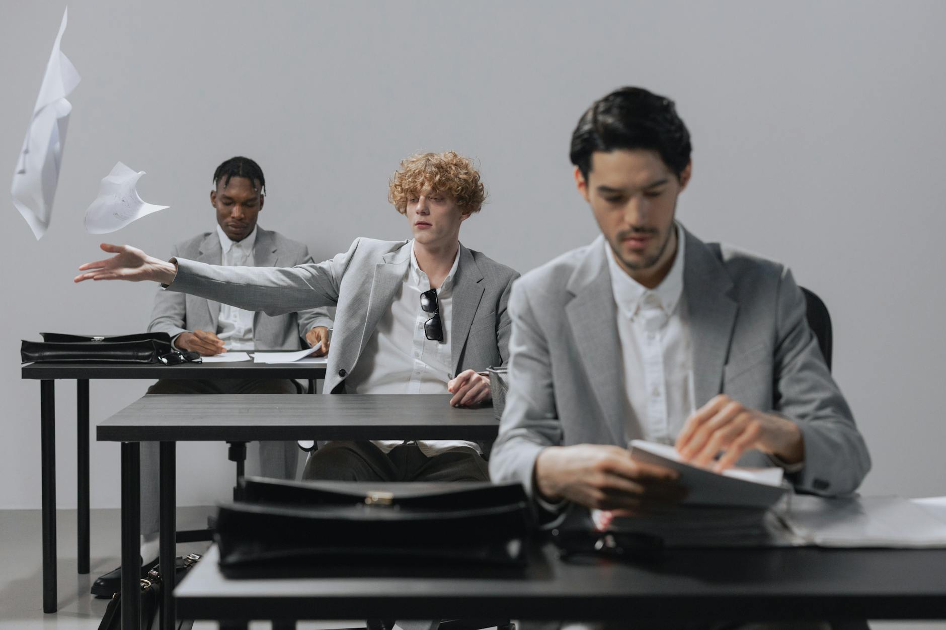 Three men in gray suits sit in an office, one tossing papers mid-air.