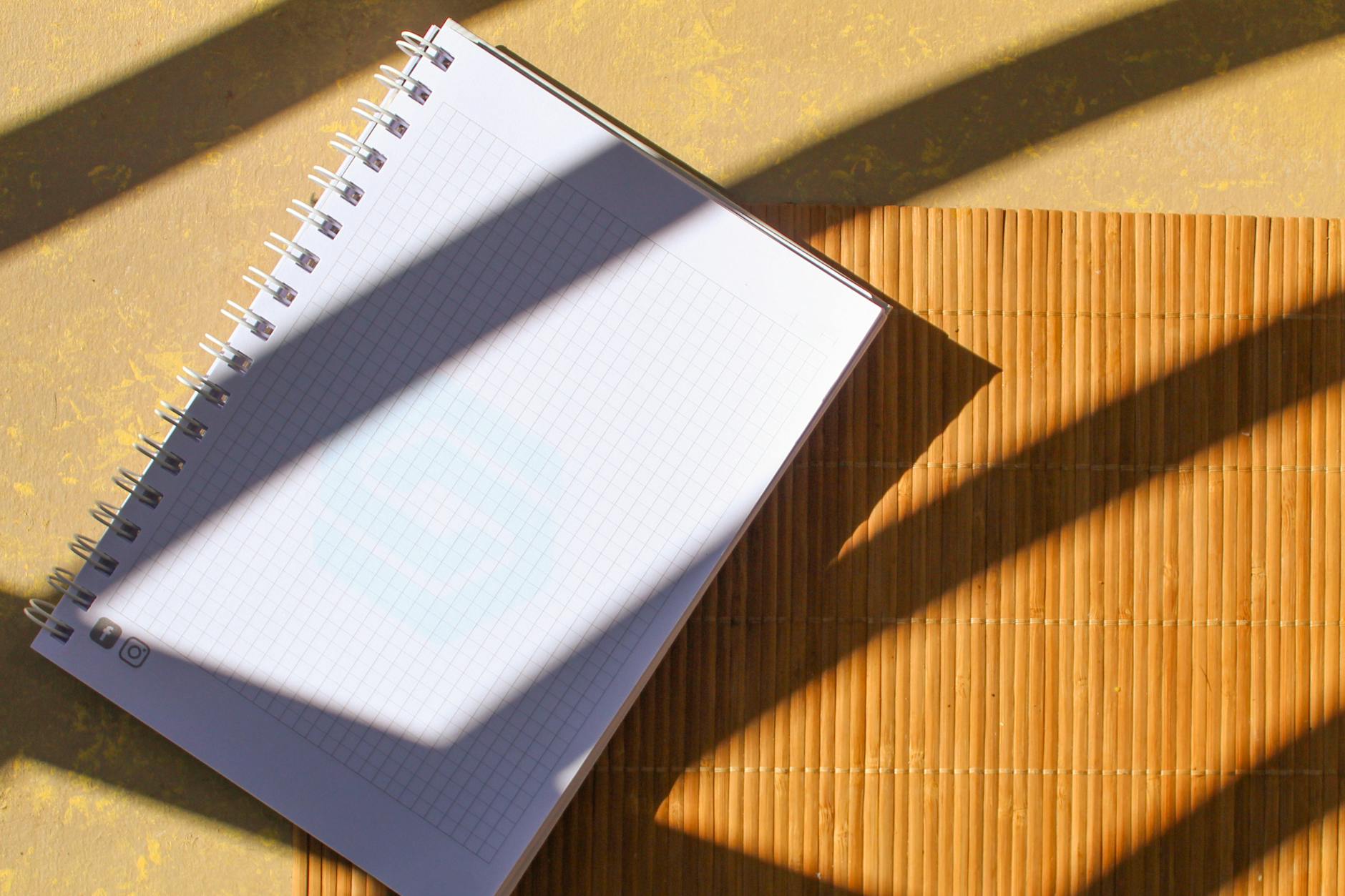 A spiral notebook placed on a bamboo mat with striking sunlight and shadow patterns.