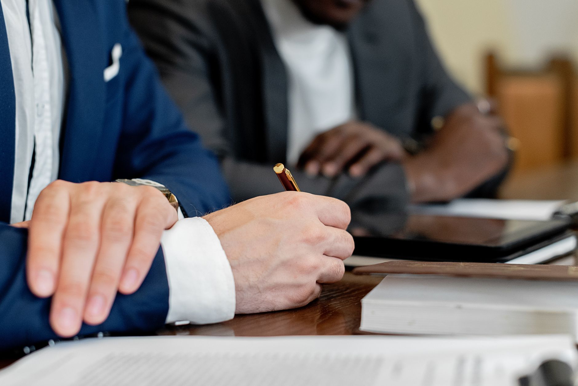 Close-up of hands writing at a business meeting. Professional attire and documents visible.