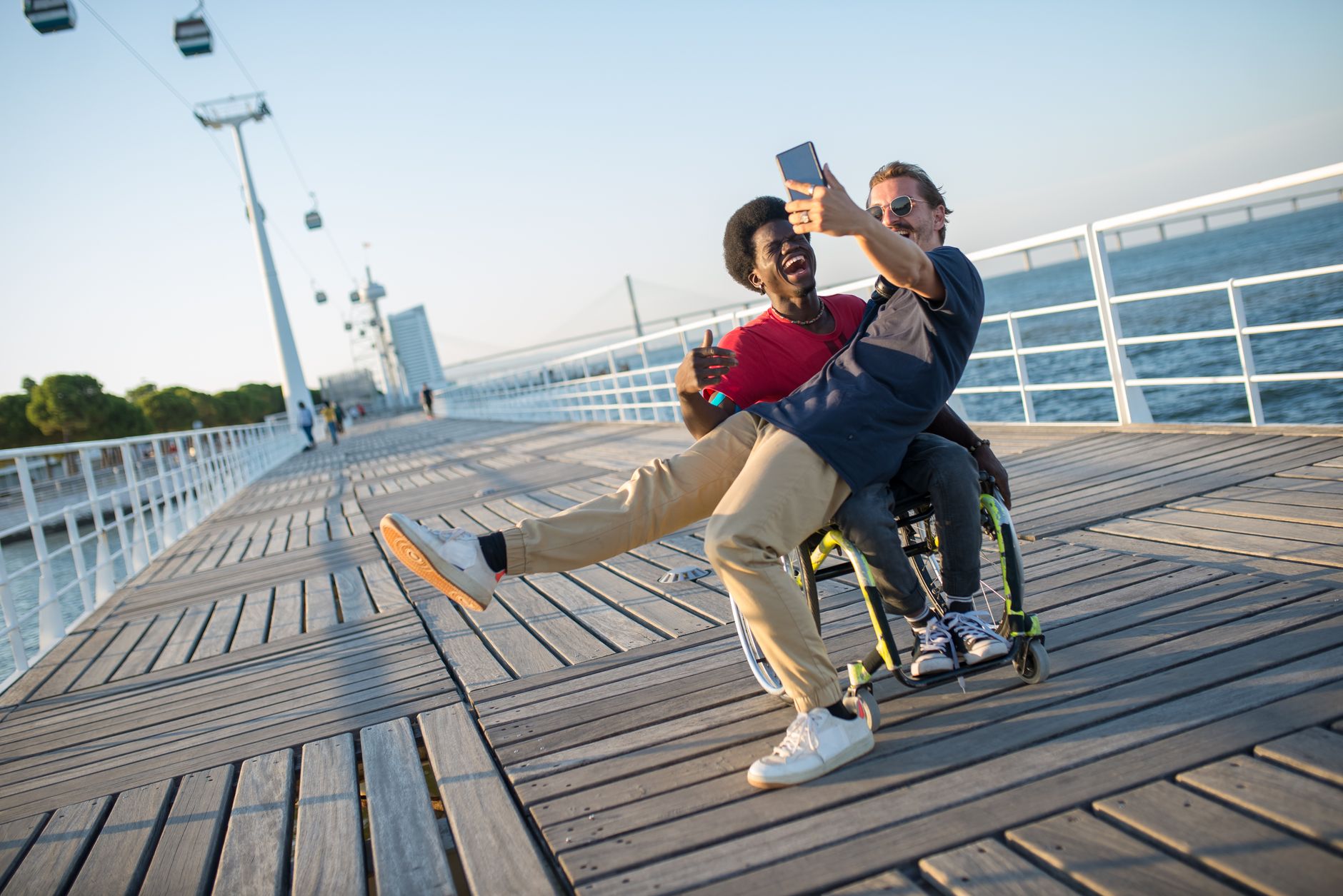 Two friends, one in a wheelchair, take a joyful selfie on a sunny boardwalk in Portugal.