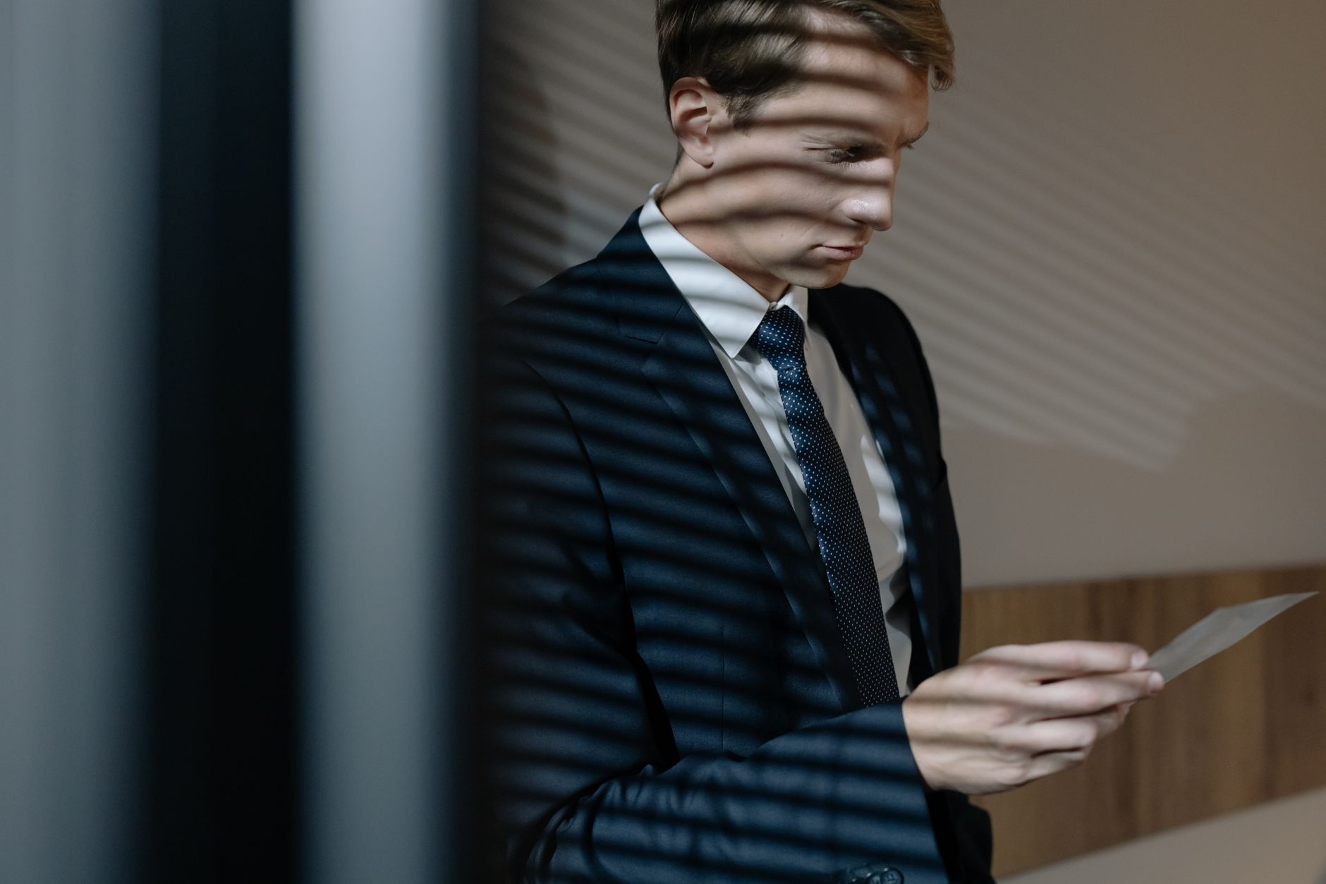Professional male in a suit examines a document with window blinds casting shadows.