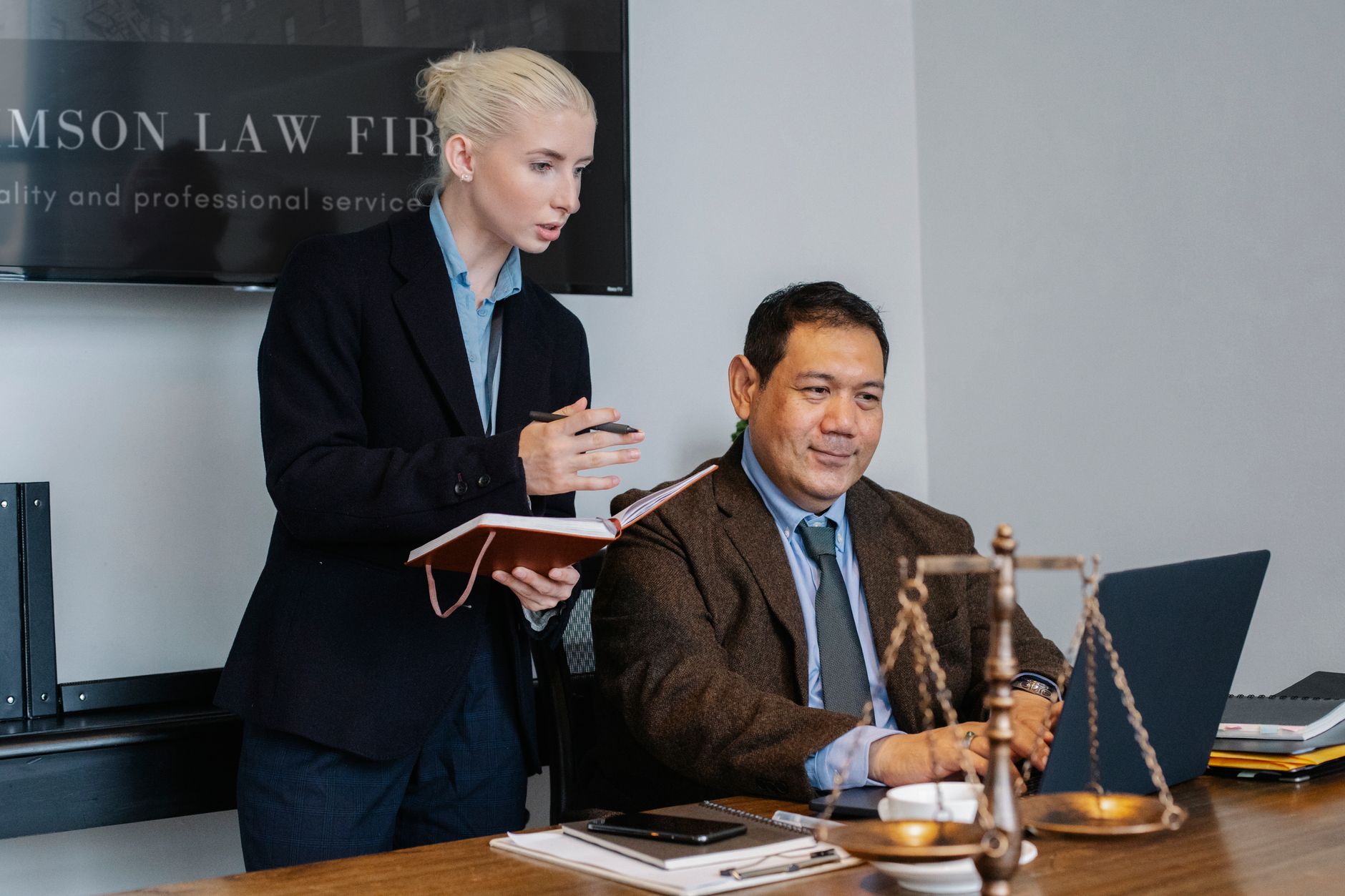 Concentrated young female assistant taking notes in planner while standing at table near middle aged ethnic male colleague typing on laptop during work in law firm