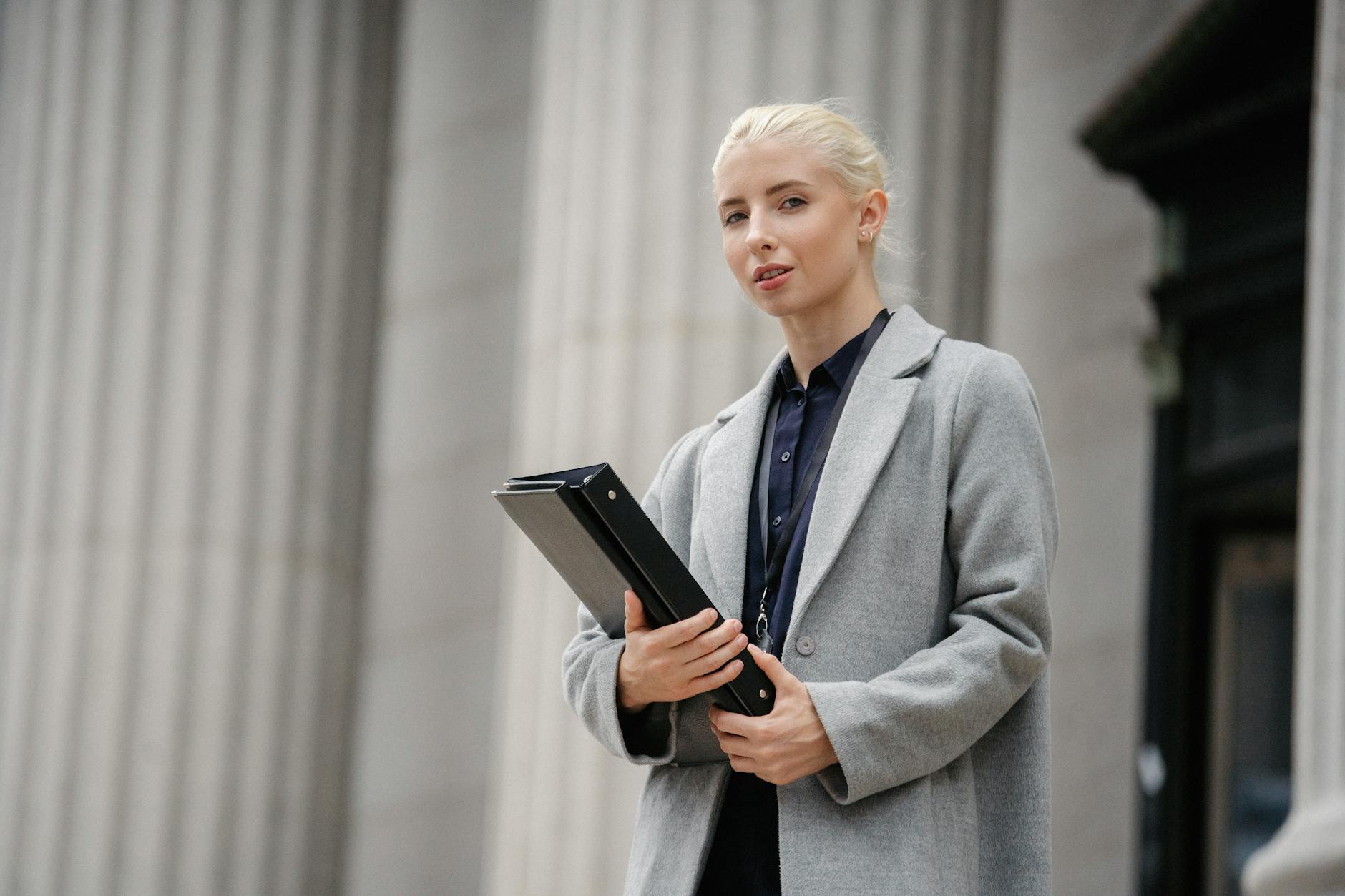 Calm young businesswoman in gray coat with black folder in hands standing outside stone building columns and looking at camera