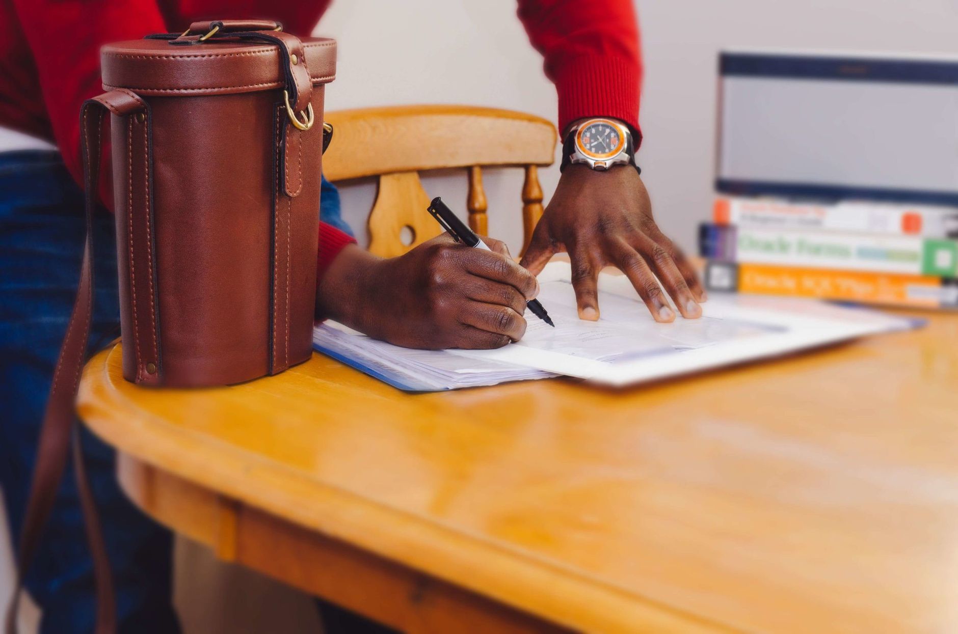 A man writing with a pen on a wooden table next to books and a leather bag.