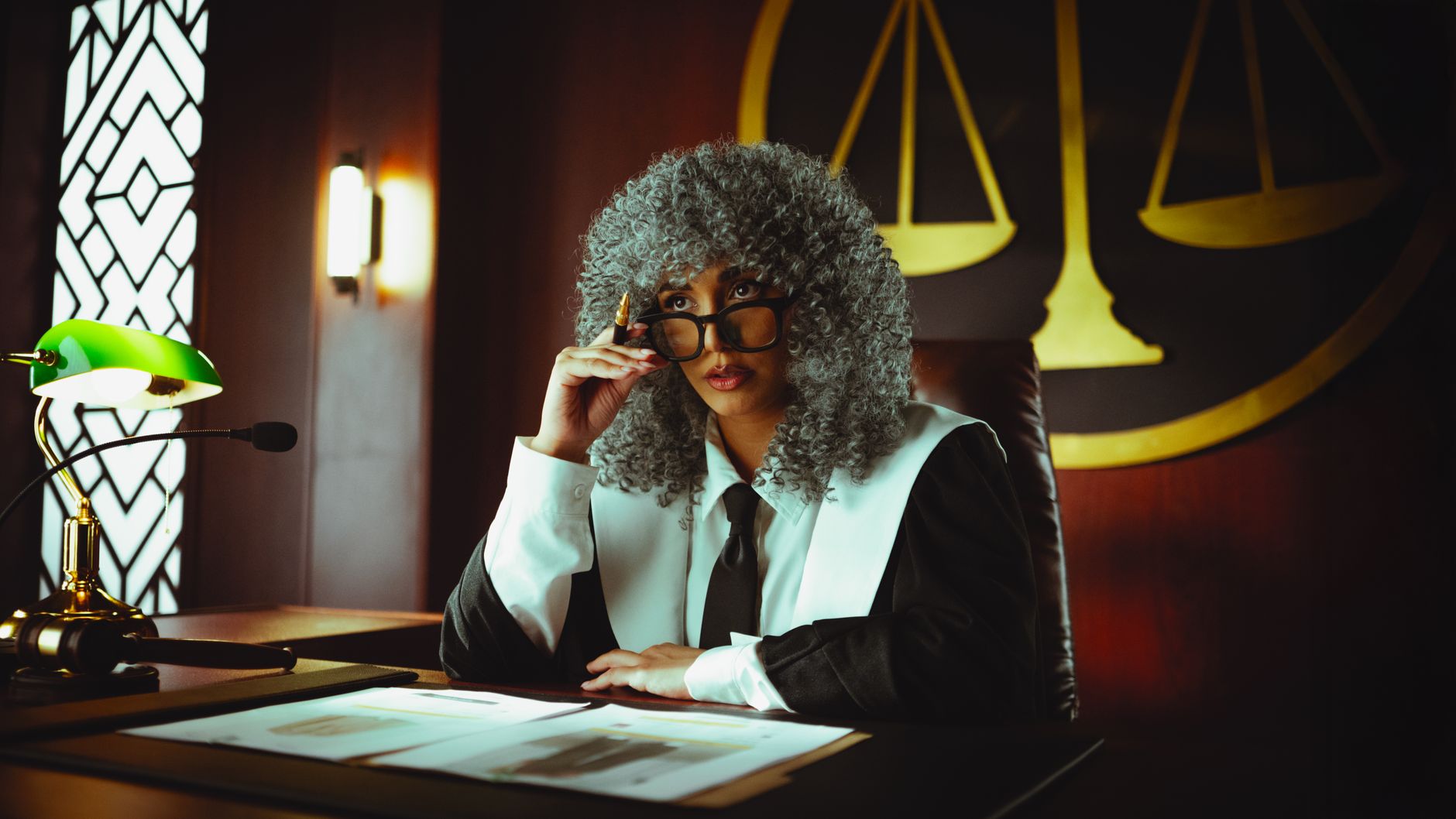 Judge in traditional attire sits at a courtroom desk with a serious expression.