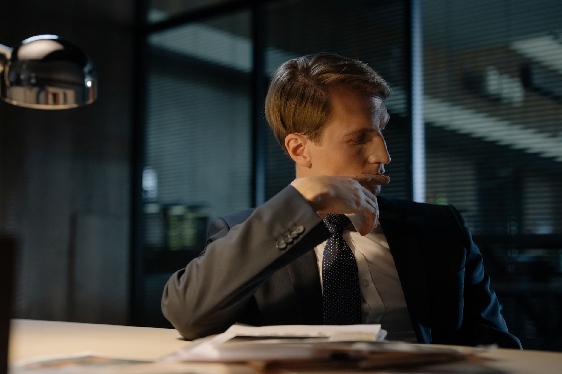 A man in a suit thoughtfully sitting at a desk in an office at night.
