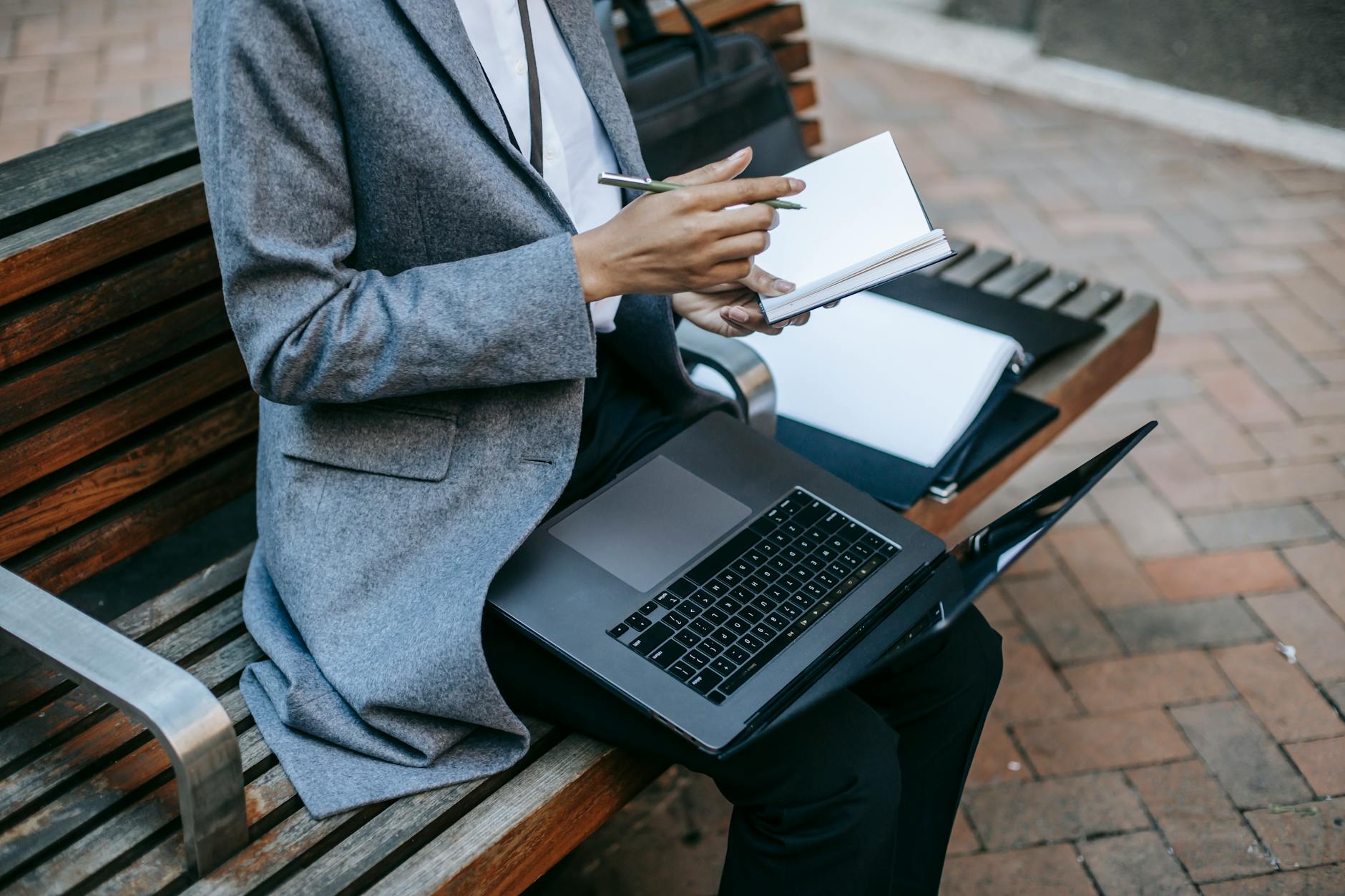 Crop faceless black businesswoman wearing gray coat sitting with laptop and papers while writing notes in planner in downtown