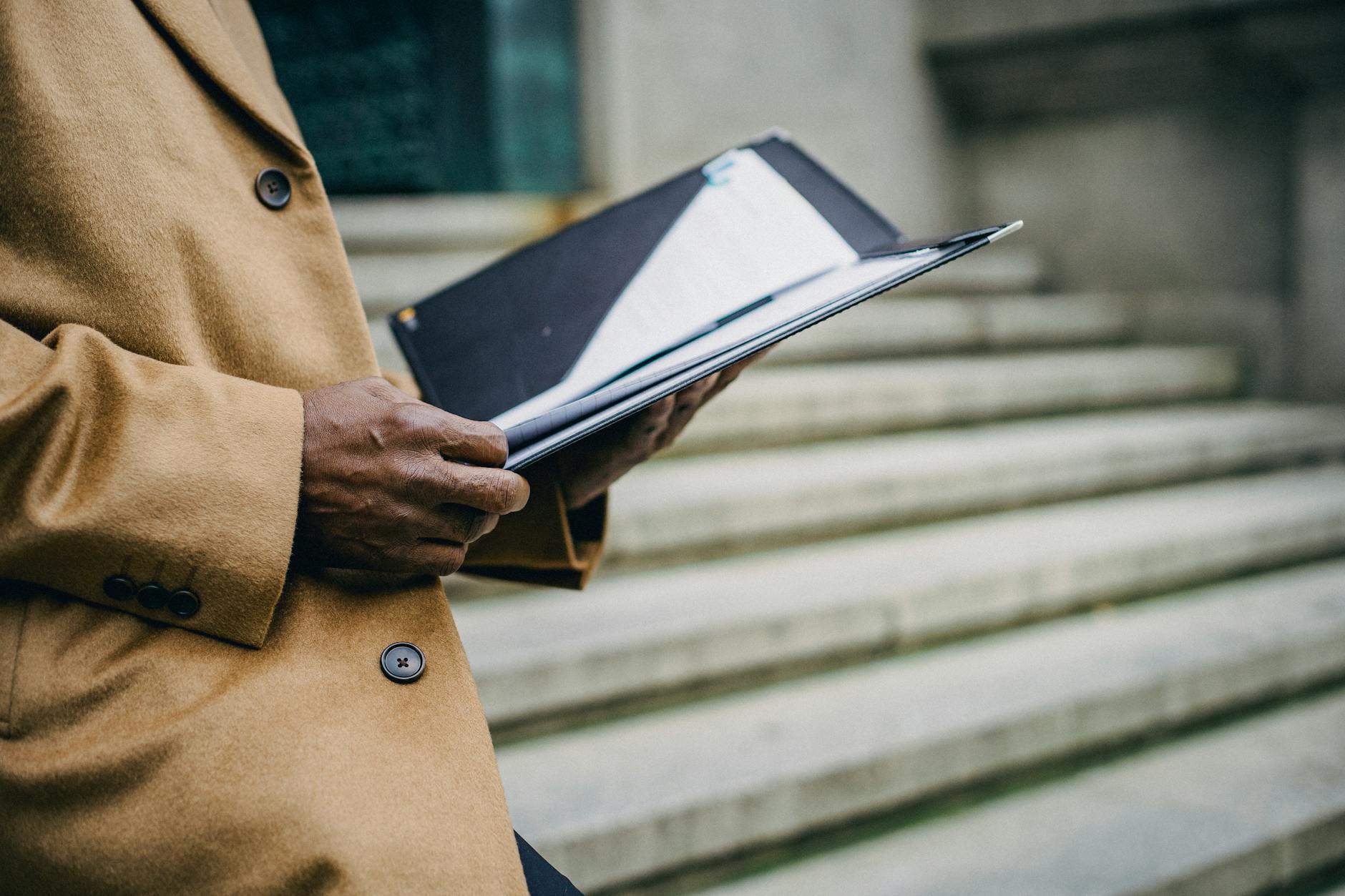 Side view of crop anonymous ethnic male executive with black folder standing on stone staircase in city