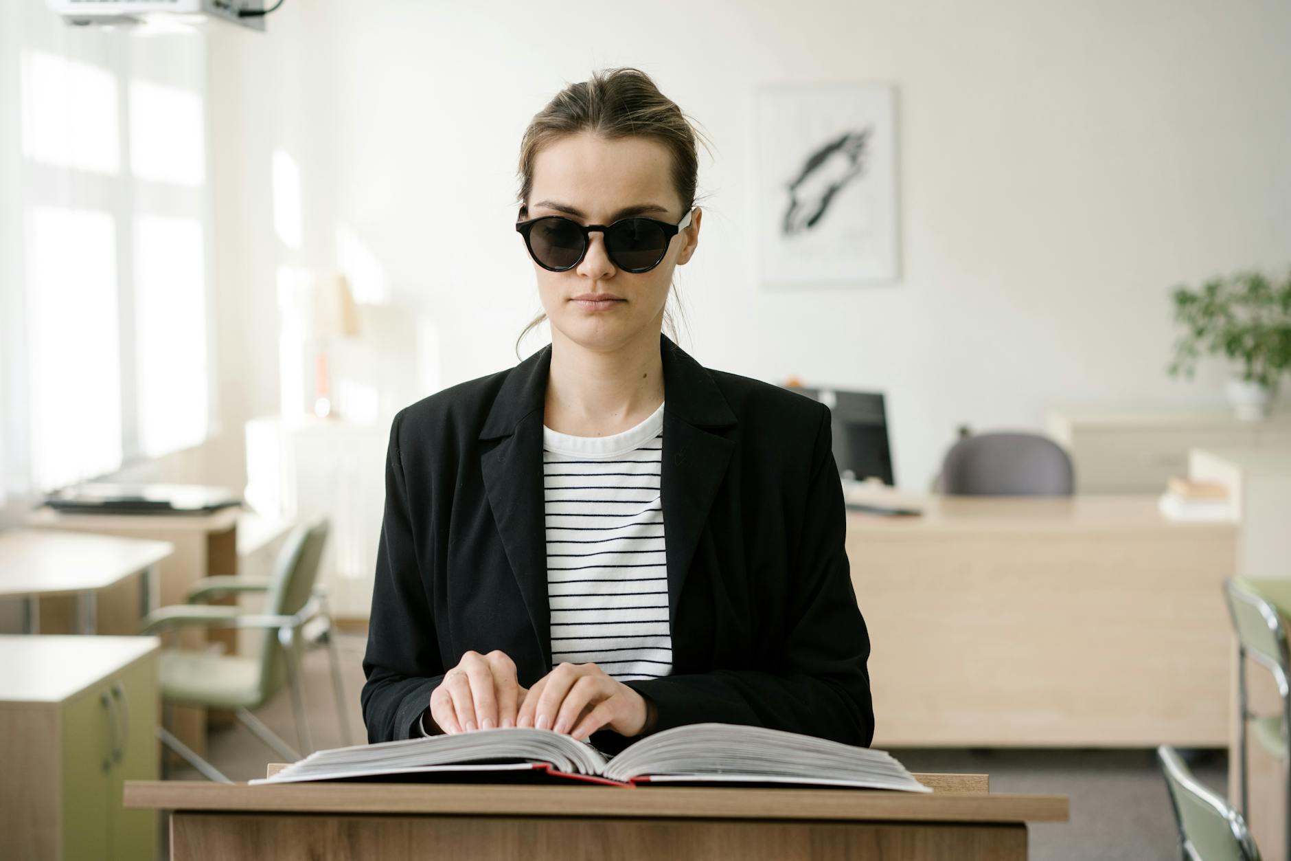 Blind woman wearing sunglasses reads a braille book at a podium in an office.