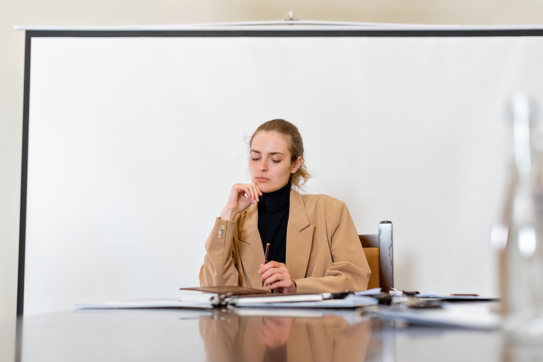 A focused young woman in a beige jacket sits at a desk with notes, deep in thought.