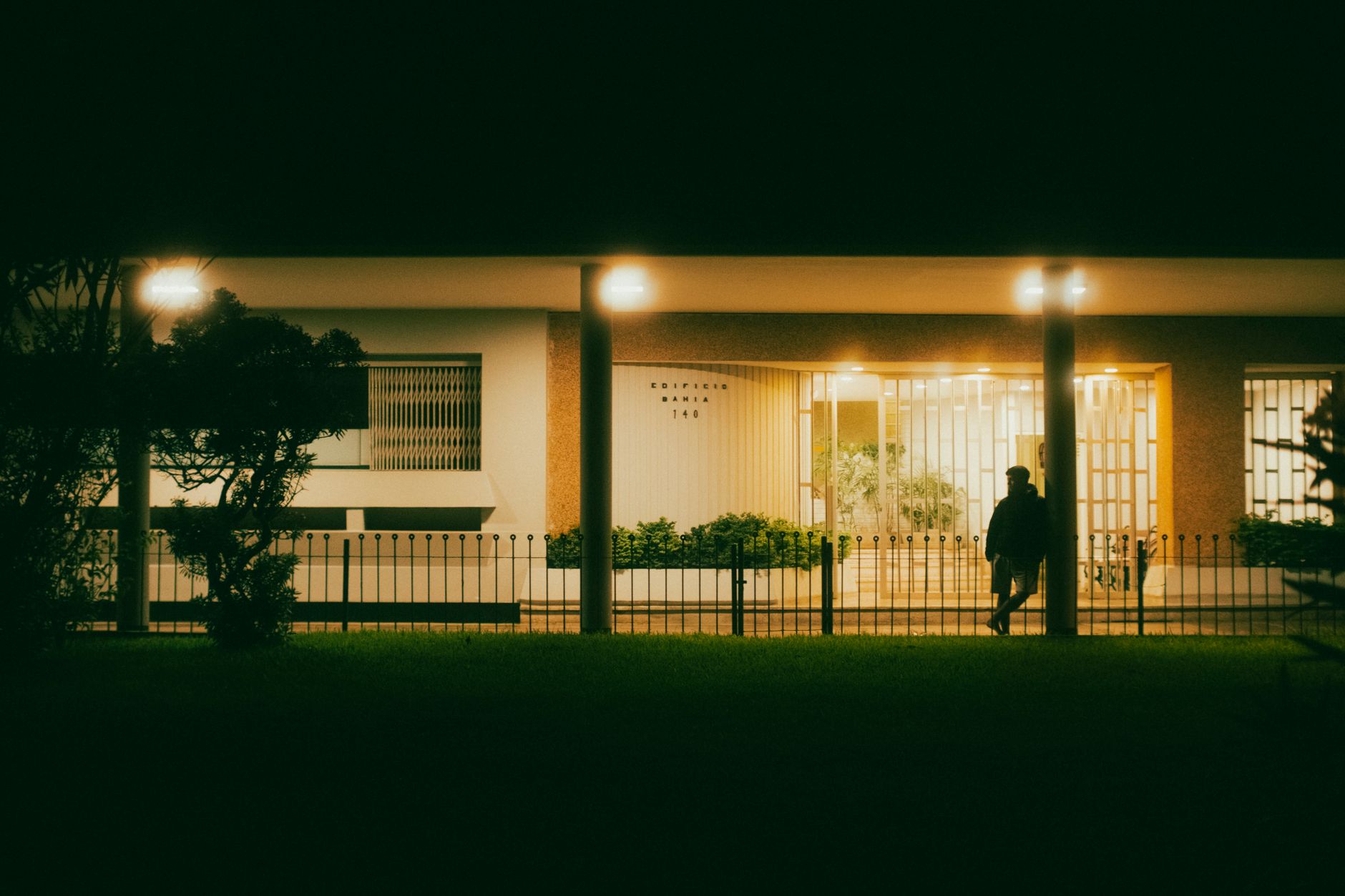 A lone figure walks by a modern building under evening lights, creating a moody scene.