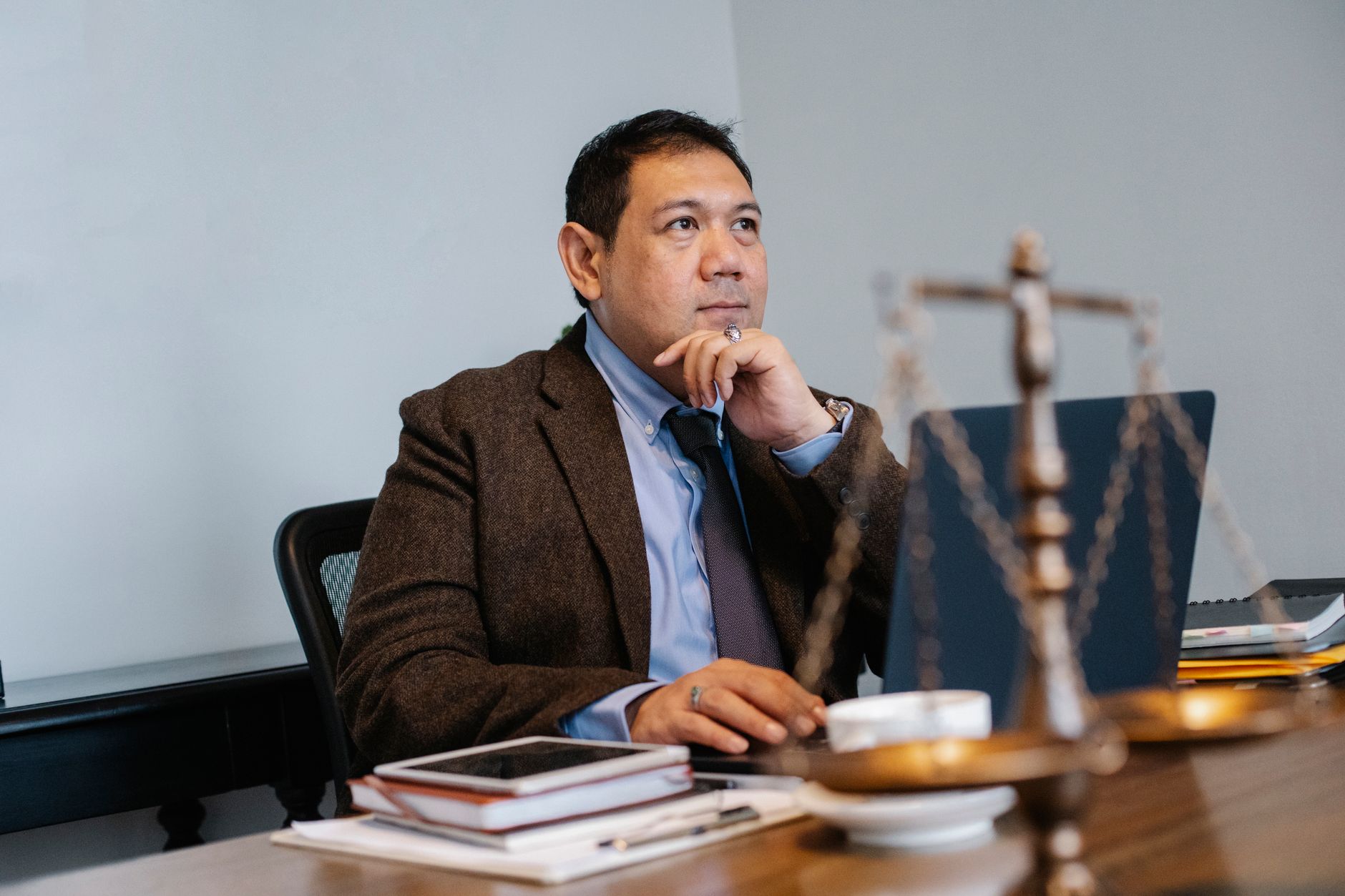 Pensive Asian male lawyer in formal suit browsing netbook while sitting at table with decorative scale of justice and touching chin in contemplation