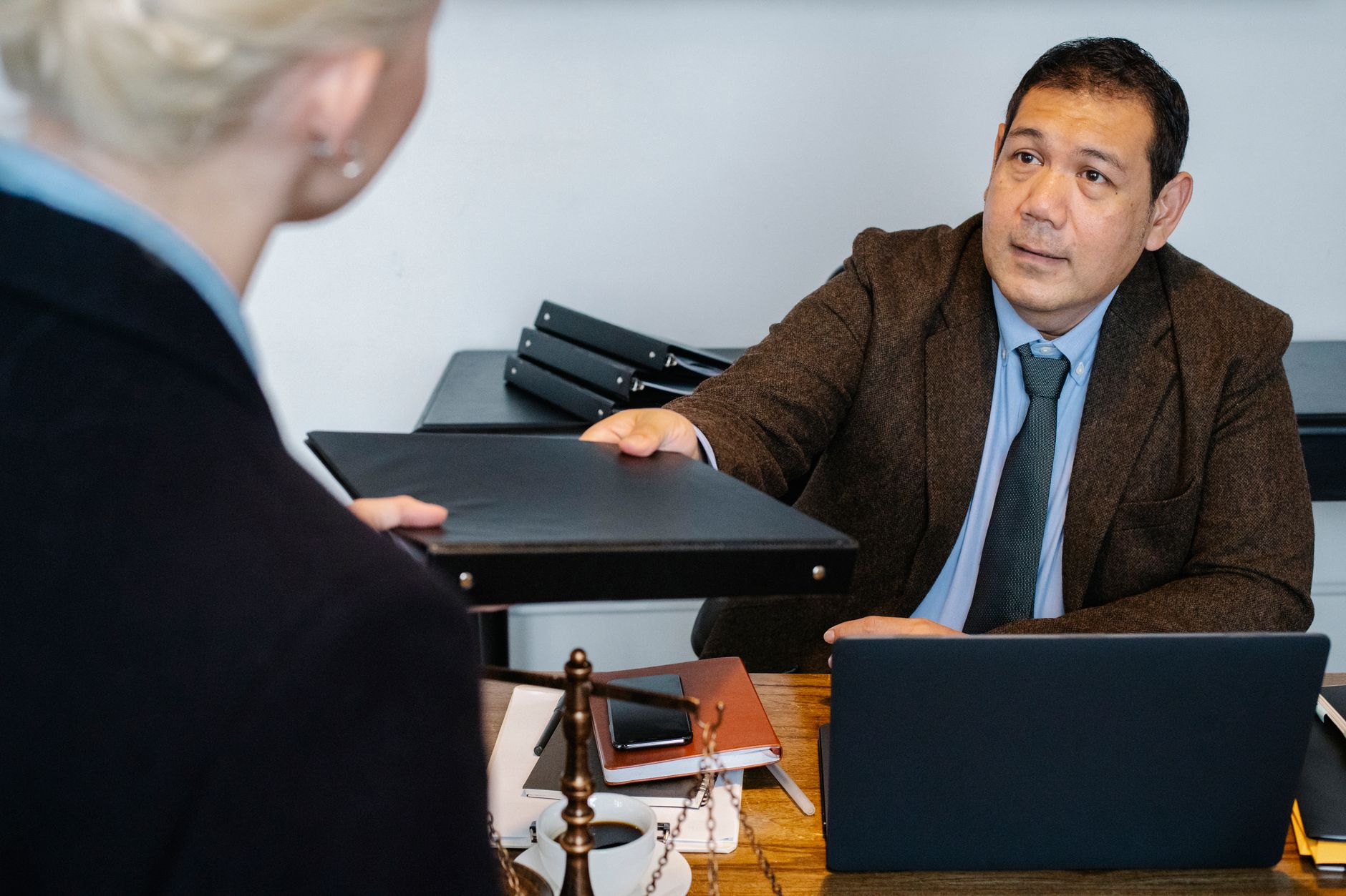 Serious middle aged ethnic male manager in suit giving document folder to unrecognizable female colleague during work at table with laptop in office