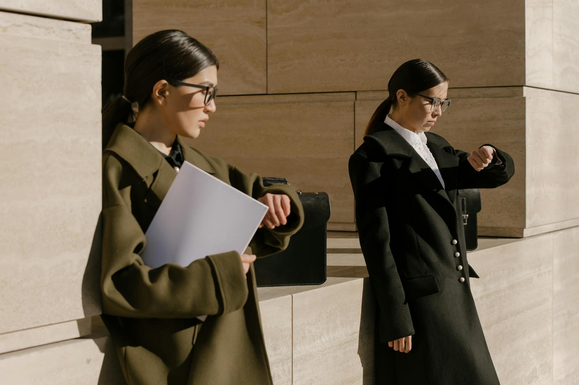 Two professional women in elegant coats and glasses, standing outdoors, checking time against modern architecture.