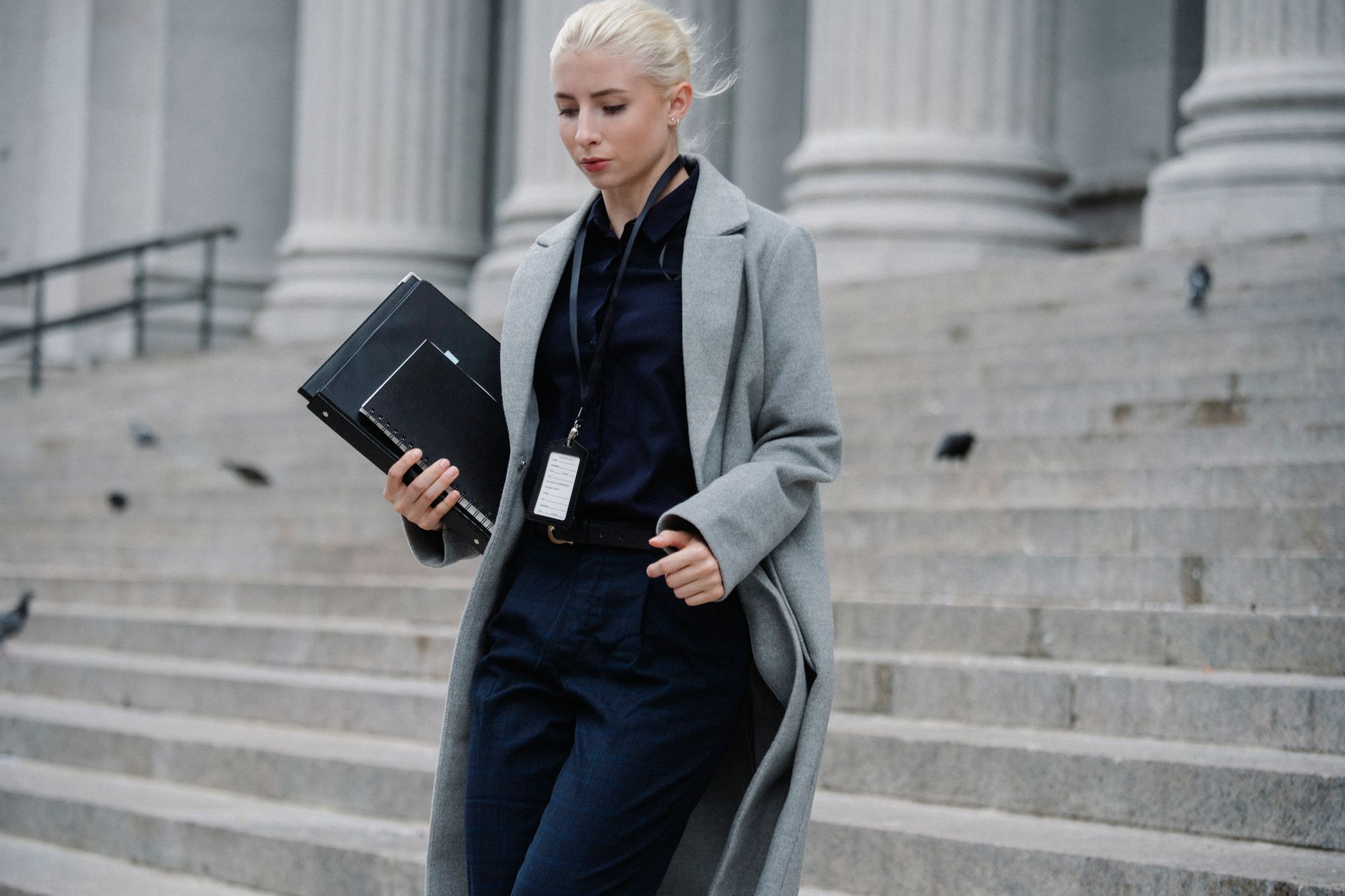 Young stylish female lawyer in formal clothes with name tag holding folders while going downstairs near stone building with columns