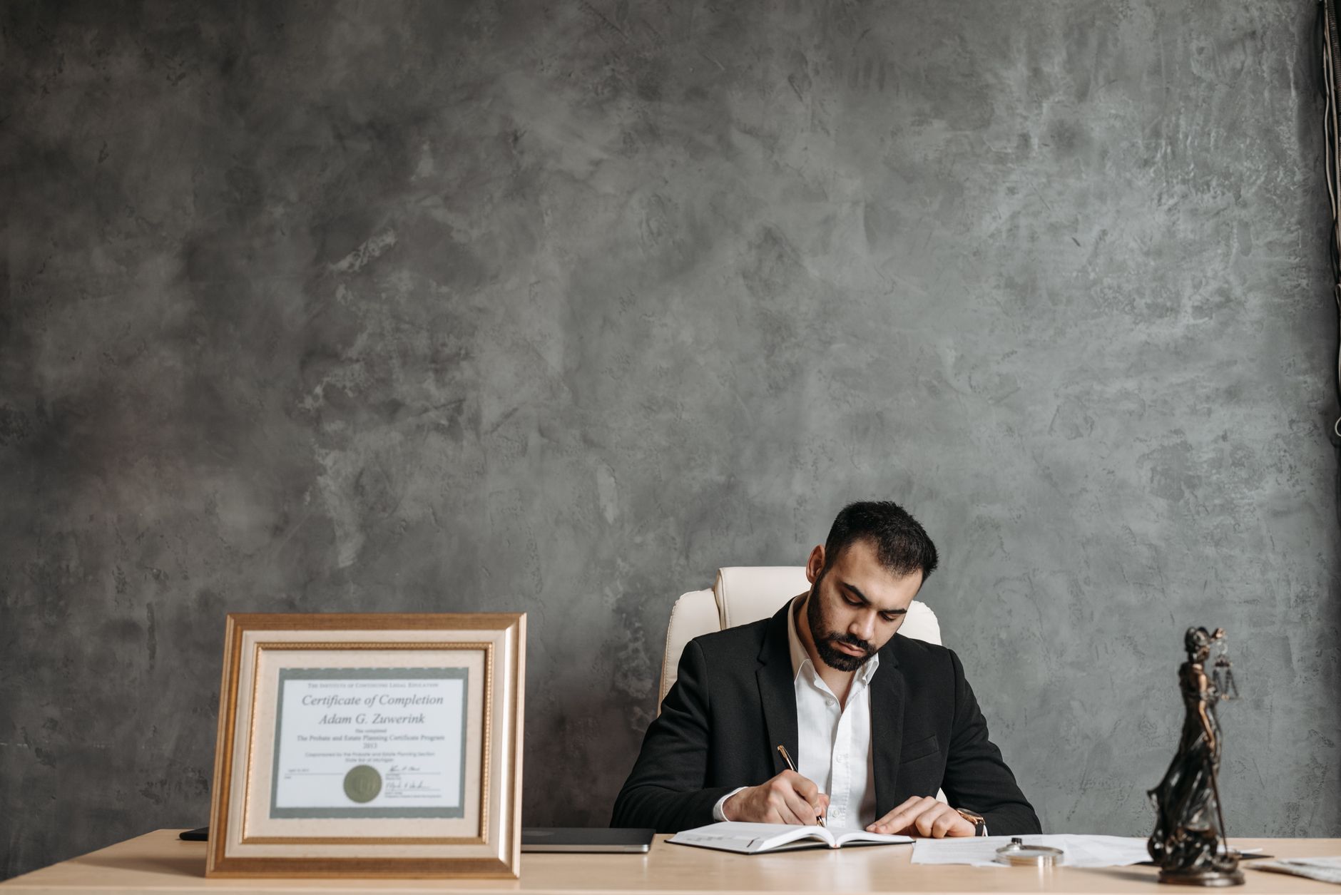 A business professional writing at a desk with a framed certificate and justice statue.