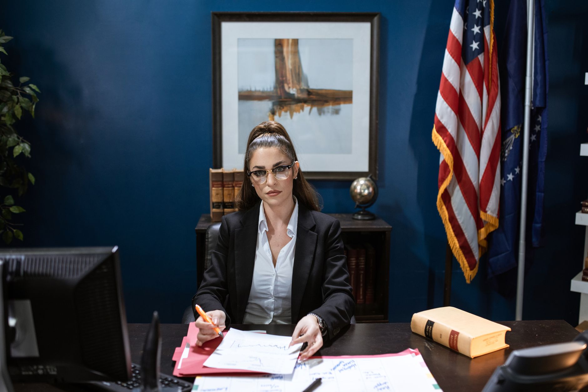 Focused woman in black blazer working on documents at desk with American flag in the background.