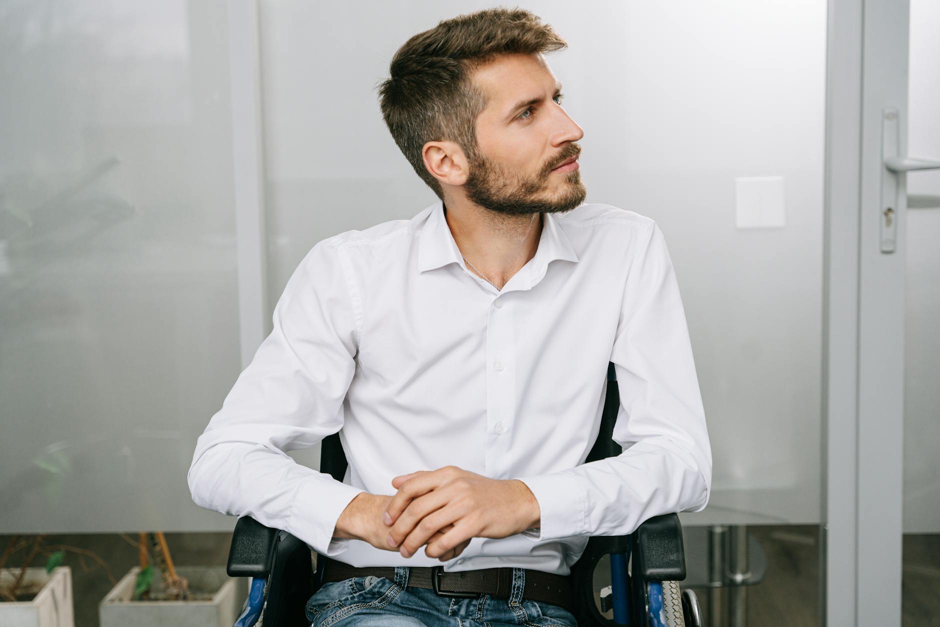 Stylish young man with a beard sitting in a wheelchair. Modern office setting, thoughtful expression.